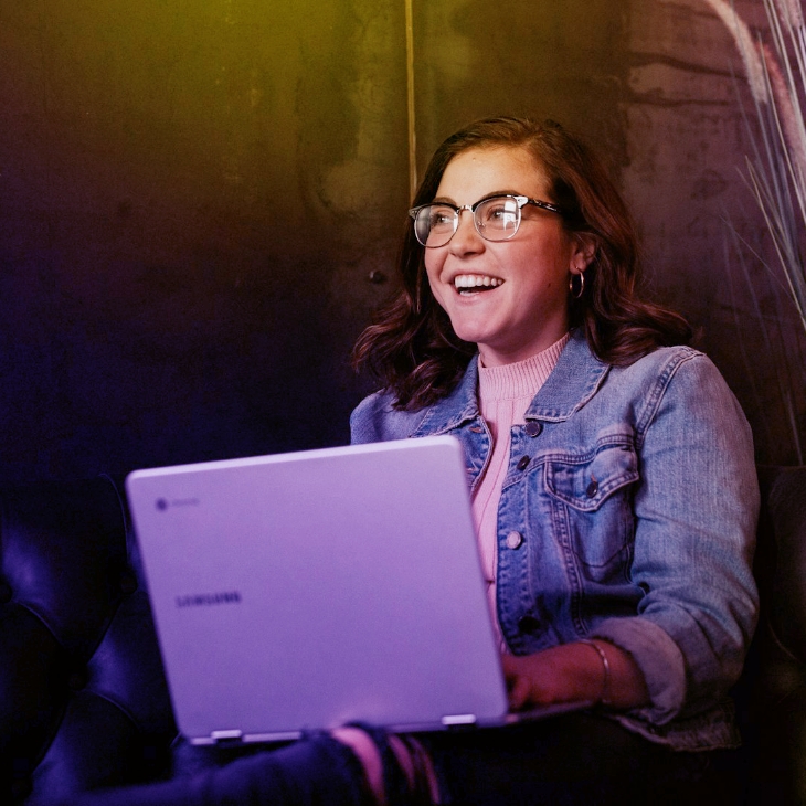 Mulher sorrindo, sentada, enquanto mexe em um notebook. Ela está olhando para o lado indicando que conversa com alguém.
