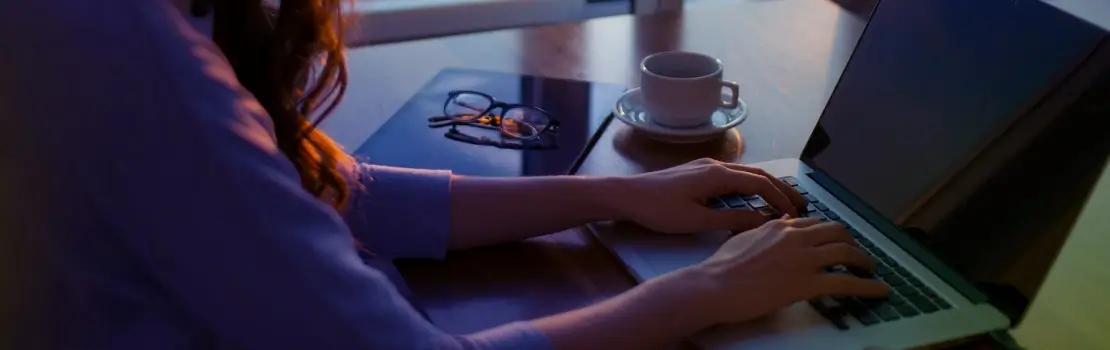 Uma pessoa sentada em frente a uma mesa com as mãos repousadas sobre o teclado do notebook. Ao lado do notebook, na mesa, há um óculos e uma xícara de café.