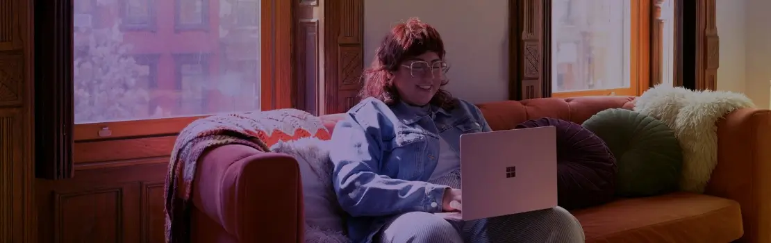 Uma mulher sentada em um sofá confortável com um notebook no colo, usando o notebook e sorrindo.